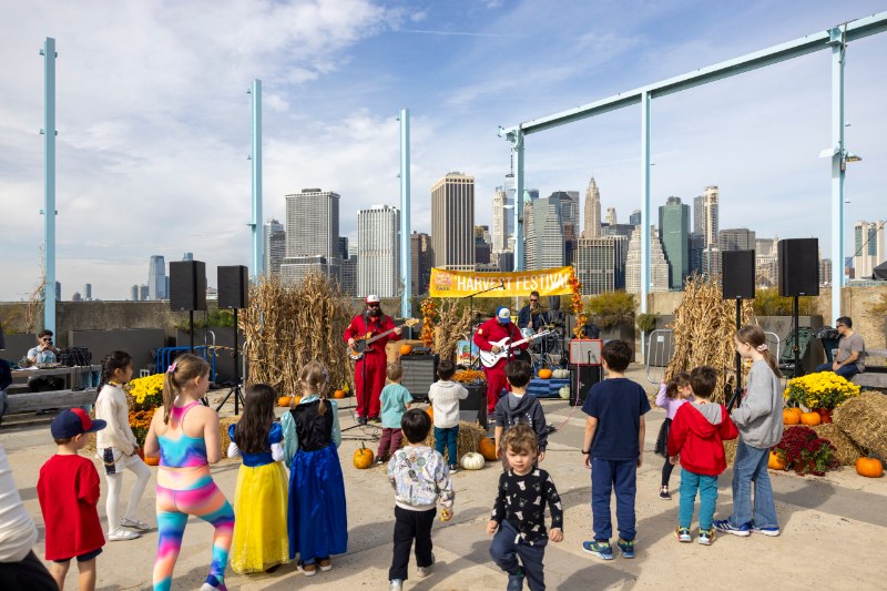 Kids in costume dance in front of a band at the Harvest Festival on Pier 6 in Brooklyn Bridge Park, with the Manhattan skyline behind