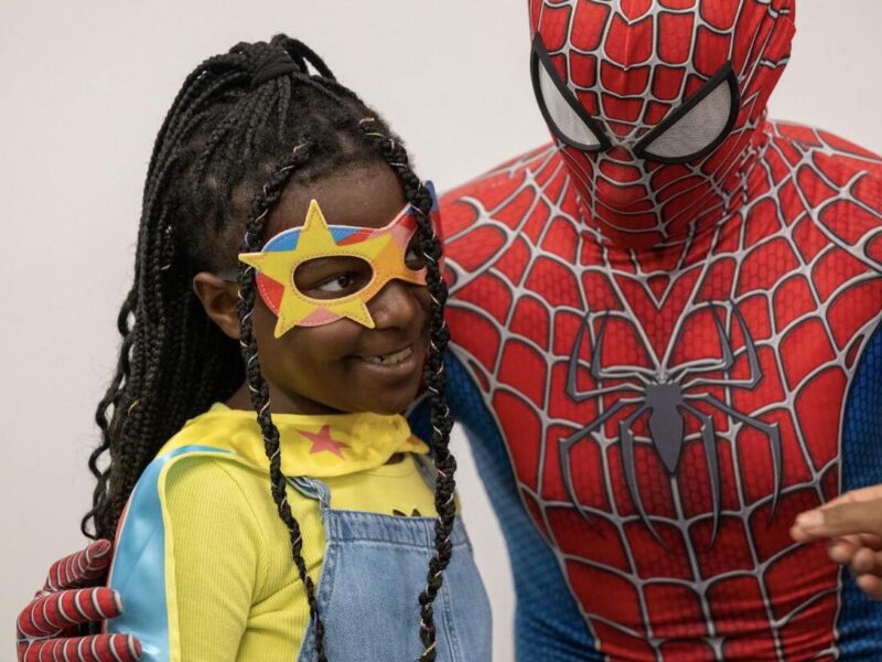 A young girl in a star mask and superhero cape smiles next to a person in a Spiderman costume at a family event