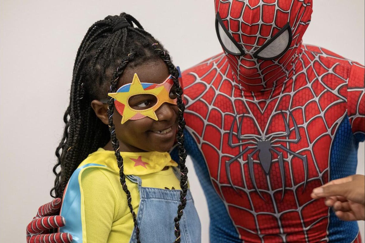 A young girl in a star mask and superhero cape smiles next to a person in a Spiderman costume at a family event