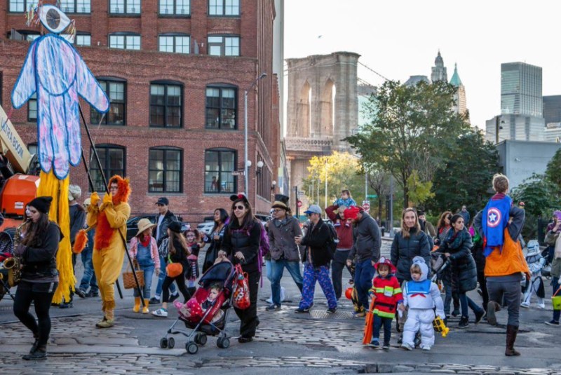 Families in costume parading past a giant puppet near the Brooklyn Bridge in DUMBO