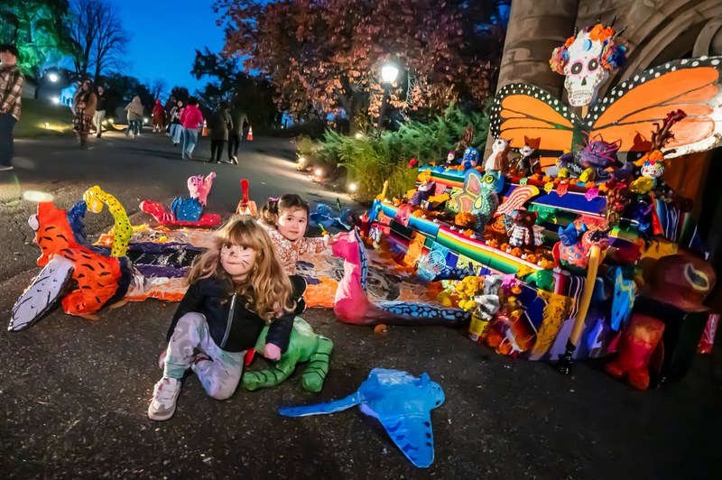 Children sit beside a colorful Day of the Dead altar with papier-mâché animals and a monarch butterfly at Green-Wood Cemetery