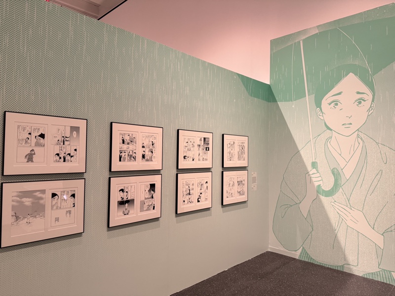 Framed manga pages and a wall mural of a woman holding an umbrella inside The Art of Manga at the Brooklyn Museum
